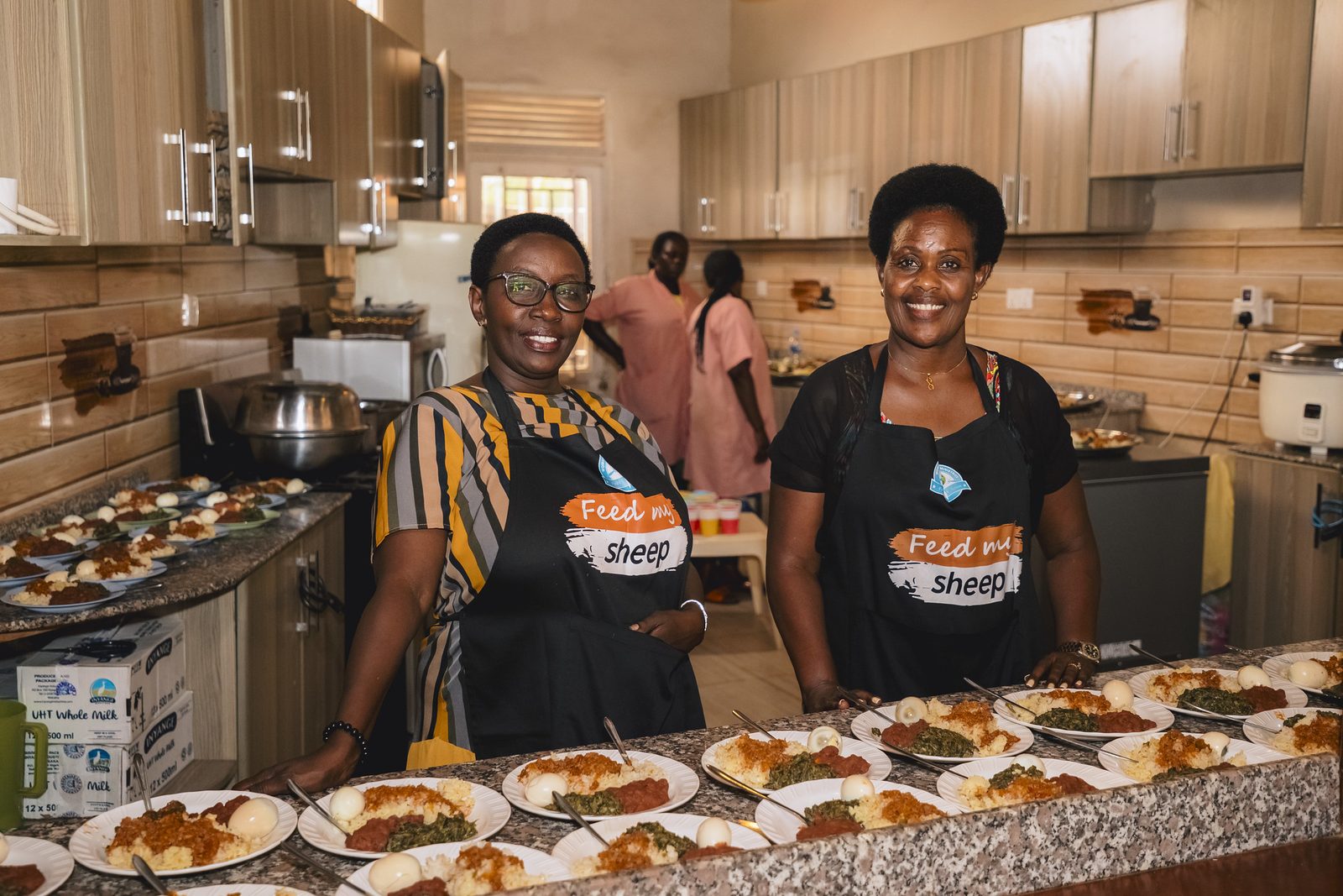 Two team members in Feed My Sheep aprons in the community kitchen, behind plates of the full food package