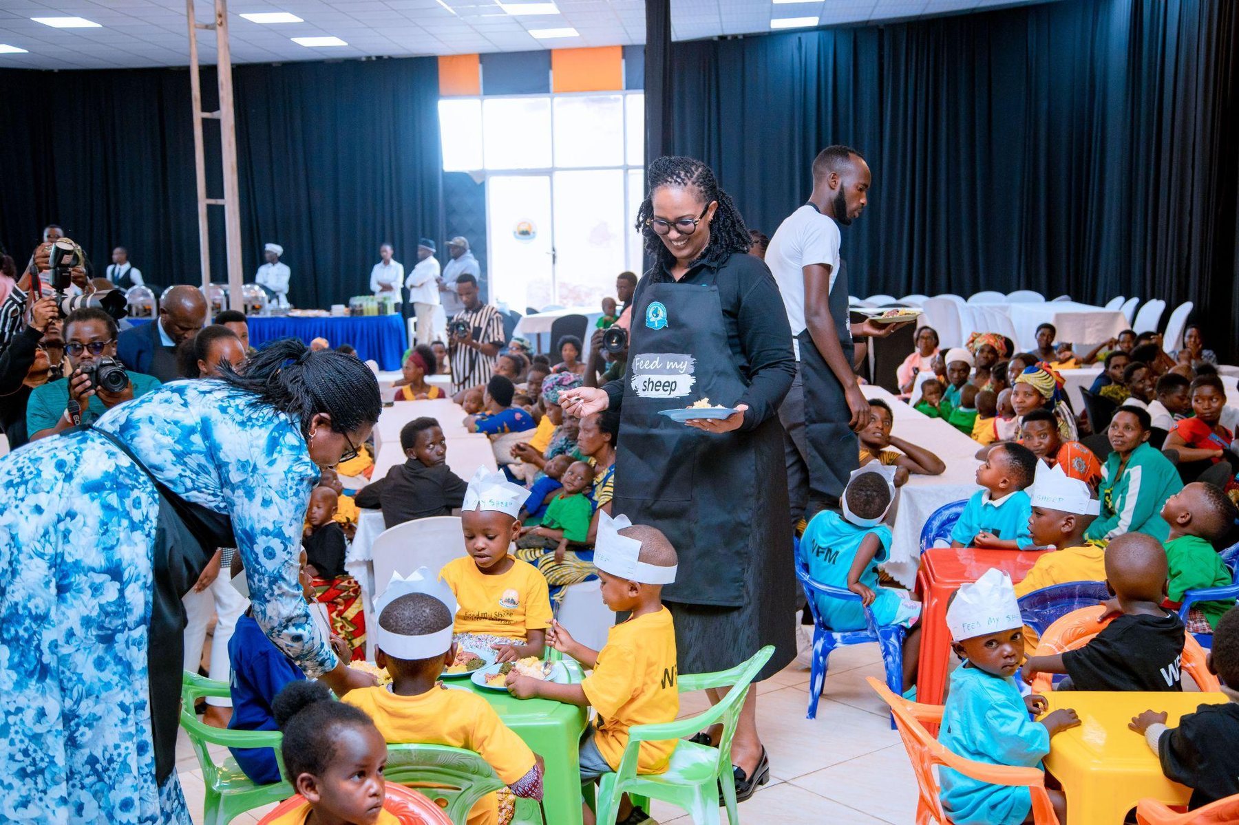 A Feed My Sheep team member in a branded apron serving a meal to children at a center celebration